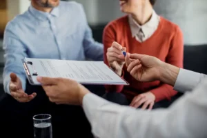 Close-up de um casal em uma mesa, com o foco na mão que segura uma caneta sobre um documento em uma prancheta, representando o ato de fazer um testamento ou de assinar um documento legal de planejamento sucessório.