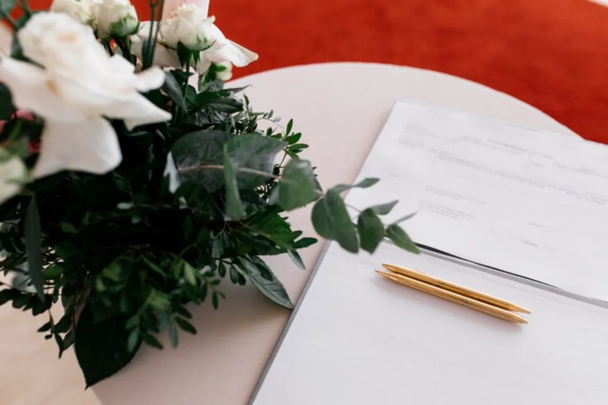 Arranjo de flores brancas ao lado de documento com canetas sobre mesa.