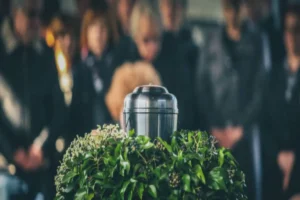 Urna funerária sobre arranjo de flores durante cerimônia de despedida, com pessoas reunidas ao fundo.